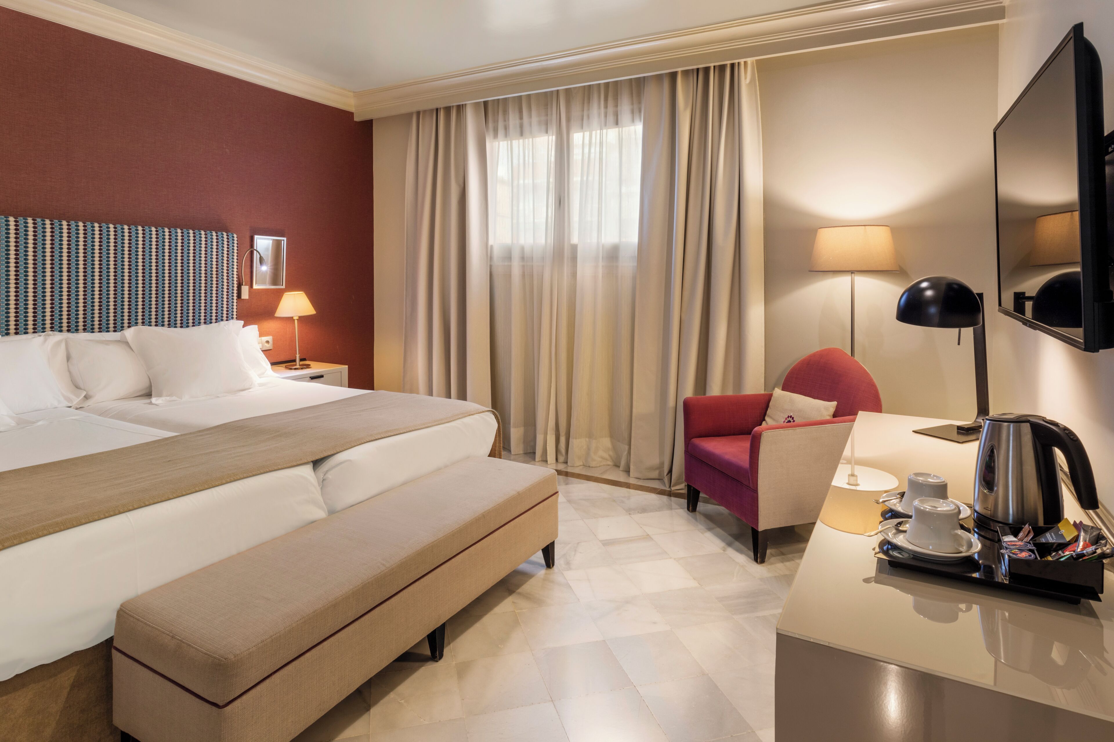 A hotel room with a large bed, a bench, a red armchair, a desk with a coffee maker, and a TV.