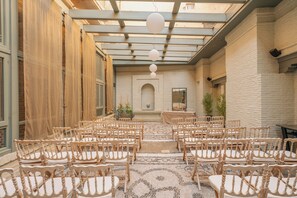 Un espace intérieur avec un mur en briques, un banc en bois et une table entourée de chaises, le tout disposé pour un événement.