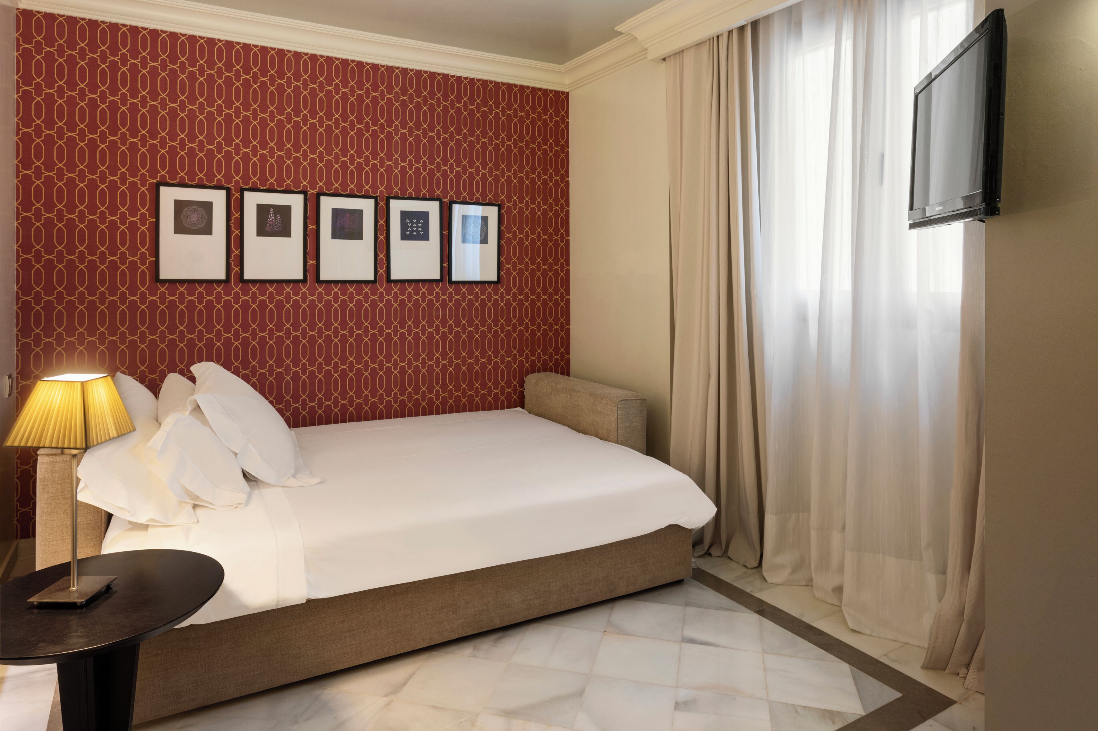 Una habitación de hotel con cama, lámpara, televisor montado en la pared y cuadros enmarcados.