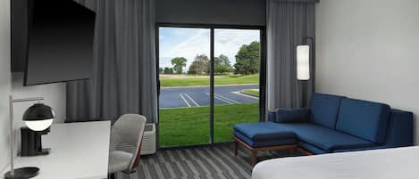 A hotel room with a bed, a desk with a chair, a TV, a window with a view of a parking lot and trees, and a lamp.