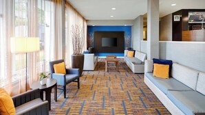 A hotel lobby with a patterned carpet, sofas, armchairs, a coffee table, and a flat-screen TV mounted on the wall. - Courtyard by Marriott Southfield (Southfield)