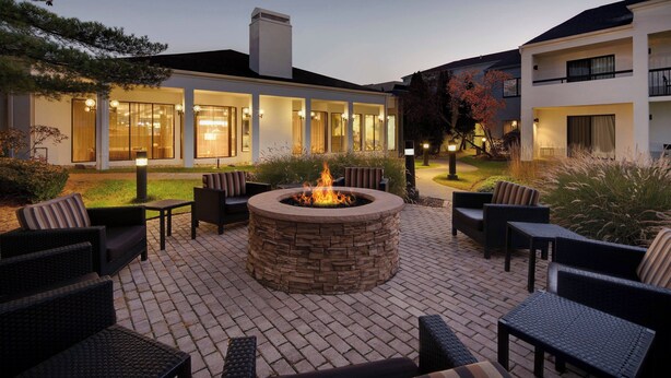 A courtyard with a stone fire pit surrounded by wicker furniture, a building with large windows and a balcony, and a well-maintained lawn. - Courtyard by Marriott Southfield (Southfield)