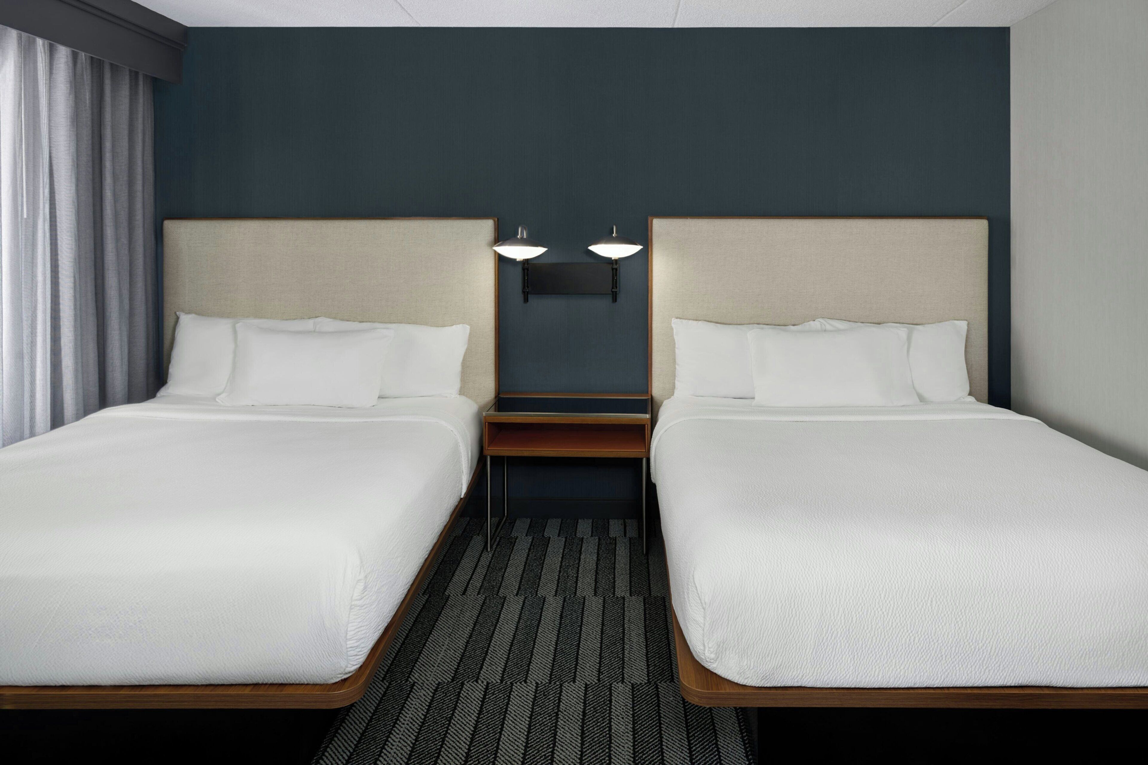 A hotel room with two single beds, a nightstand, a lamp, and a window with curtains.