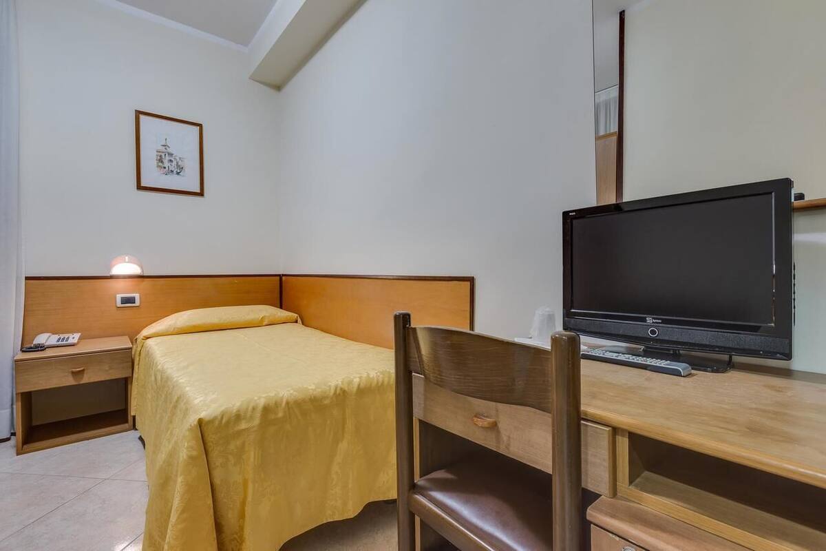 Una camera d'hotel con un letto, una scrivania con computer e tastiera, una sedia, un comodino con il telefono e un quadro appeso al muro.