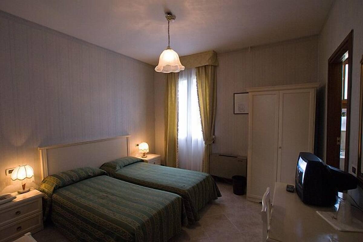 Camera d'albergo con due letti singoli, una scrivania con un computer, una sedia, una lampada e una finestra con tende.