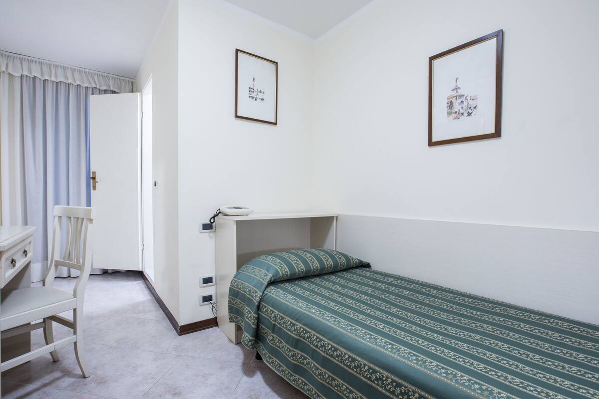 Una stanza piccola e pulita con un letto singolo, una scrivania e una sedia. Due quadri appesi al muro.