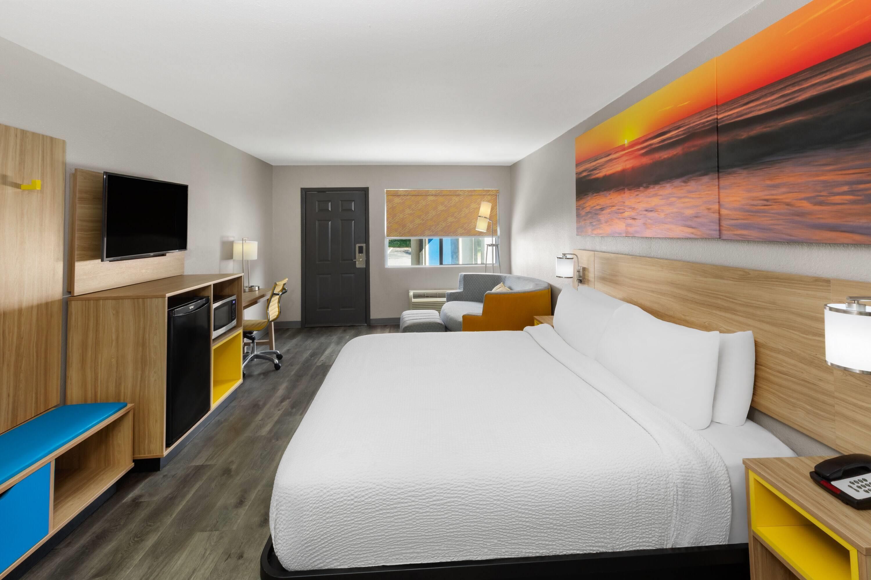 Quarto de hotel com cama, escrivaninha, televisão e uma pintura do pôr do sol.