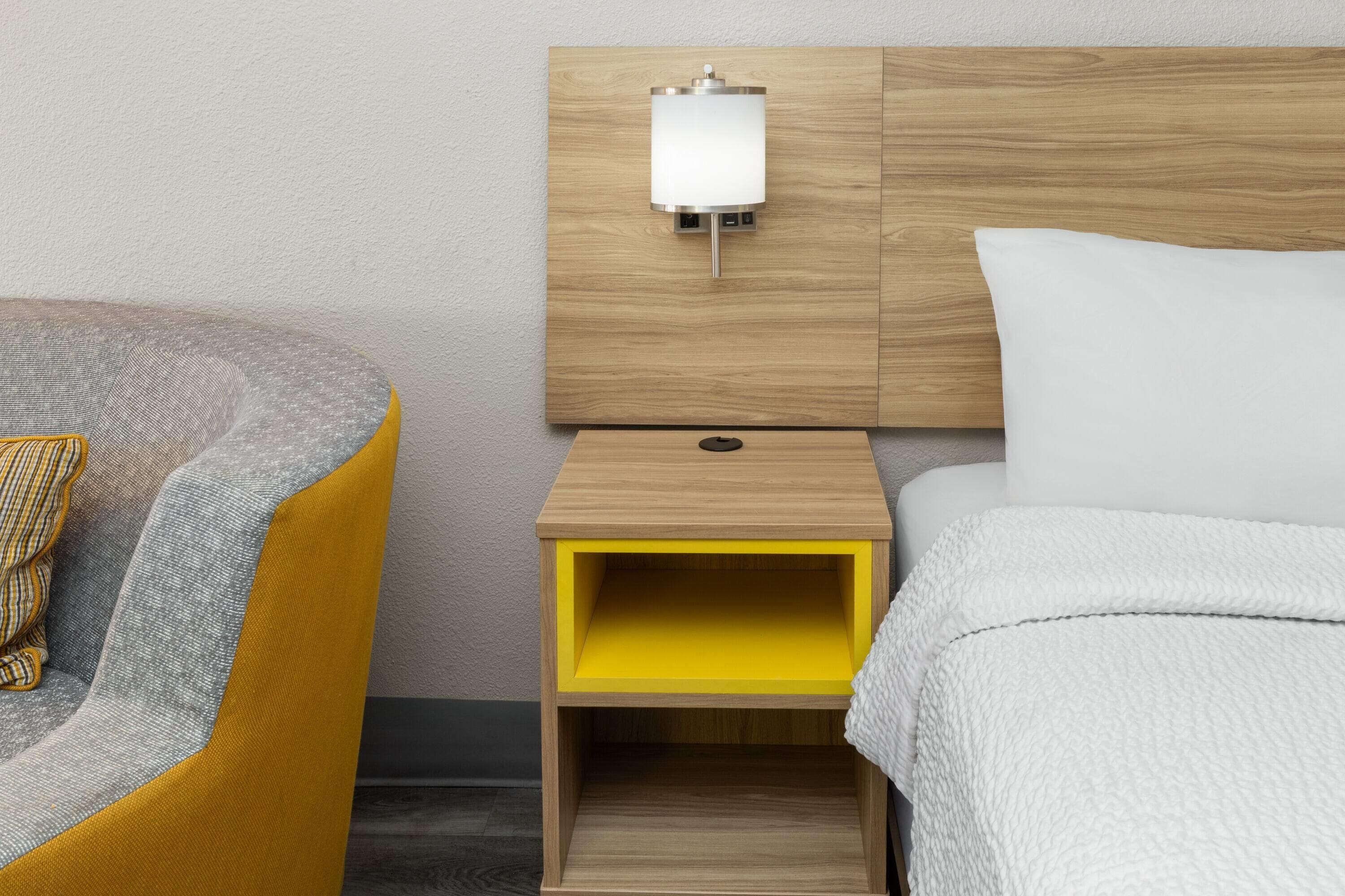 Una habitación de hotel con una cama, una mesita de noche con lámpara, una silla y un pequeño armario amarillo.