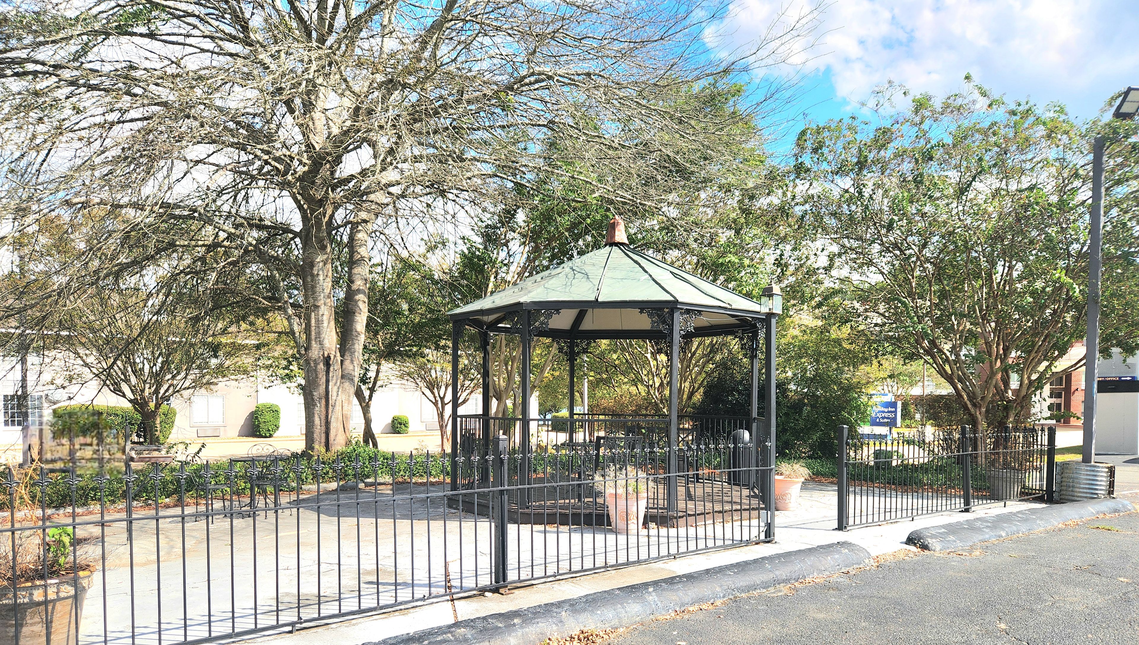 A gazebo in a park with a paved walkway and a metal fence.