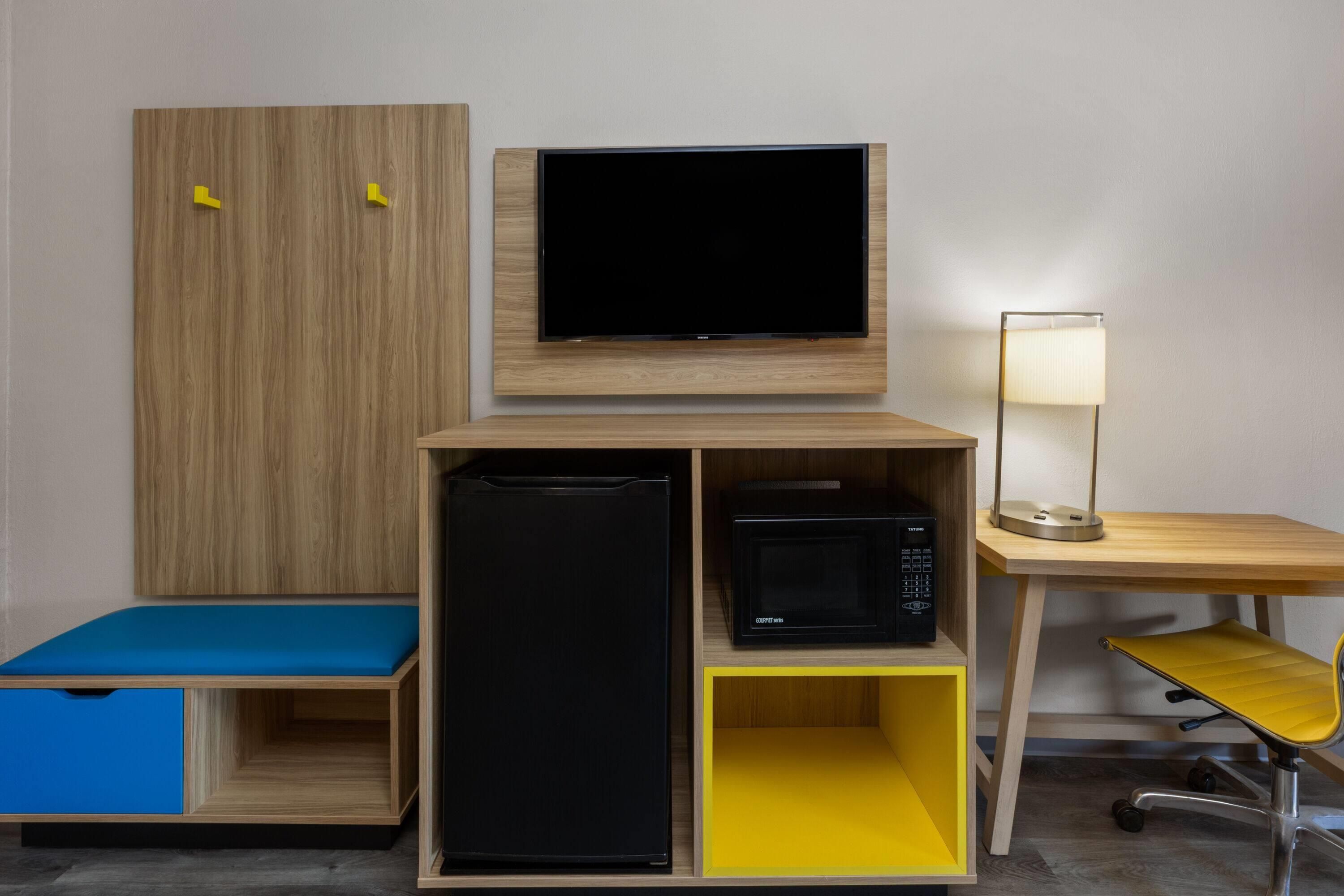 Quarto compacto com televisão na parede, escrivaninha com luminária, micro-ondas e um pequeno espaço de estar com um almofada azul.