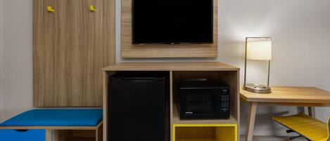 Une chambre compacte avec un téléviseur fixé au mur, un bureau avec une lampe, un four à micro-ondes et un petit coin salon avec un coussin bleu.