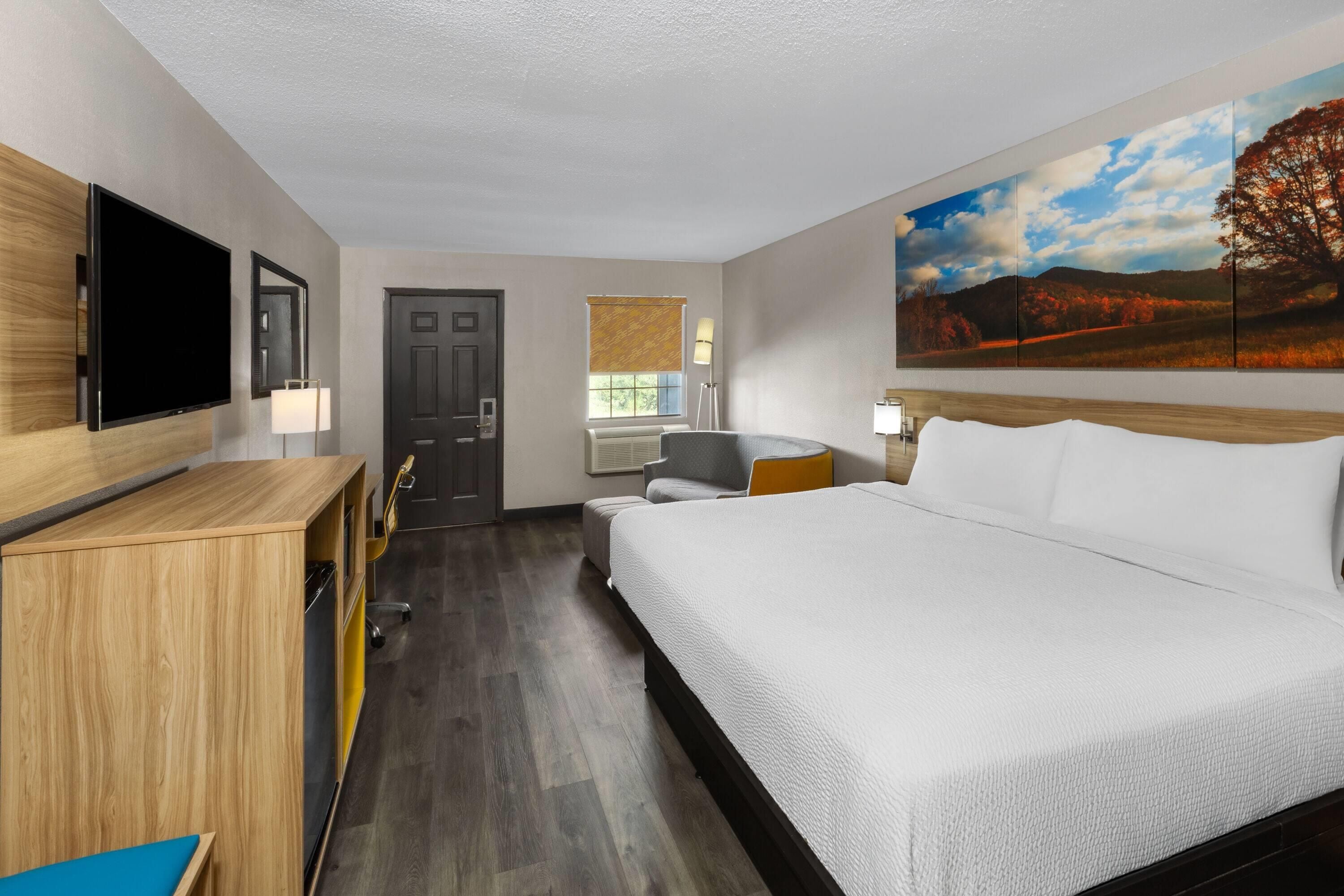 Habitación de hotel con cama, escritorio, silla, televisor, ventana con persianas y un cuadro en la pared.