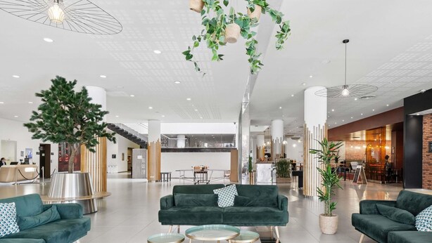 Un hall d’entrée moderne avec des canapés verts, un grand arbre et un bar à café.