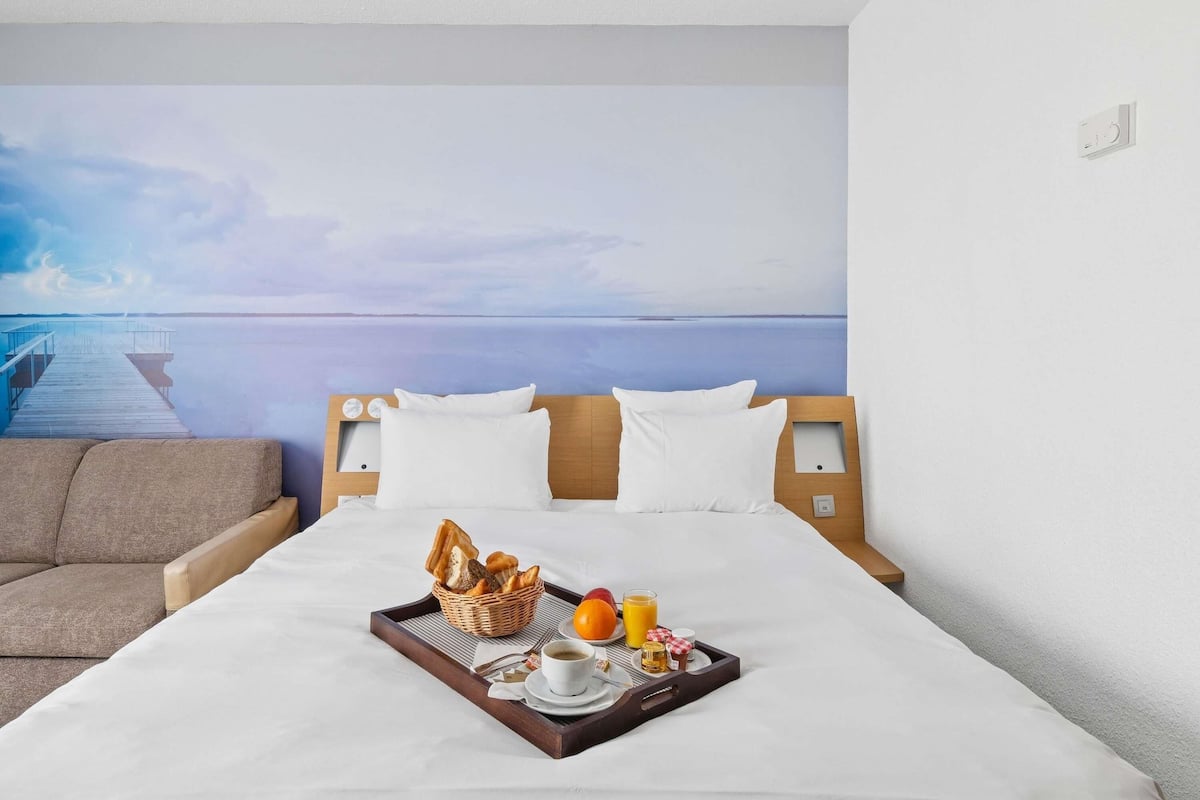Una habitación de hotel con cama, bandeja de desayuno, cabecera de madera y un mural en la pared que muestra un muelle y agua.