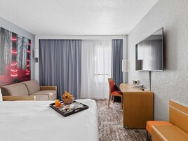 Habitación de hotel con cama, escritorio, silla, televisor y un amplio ventanal con cortinas.