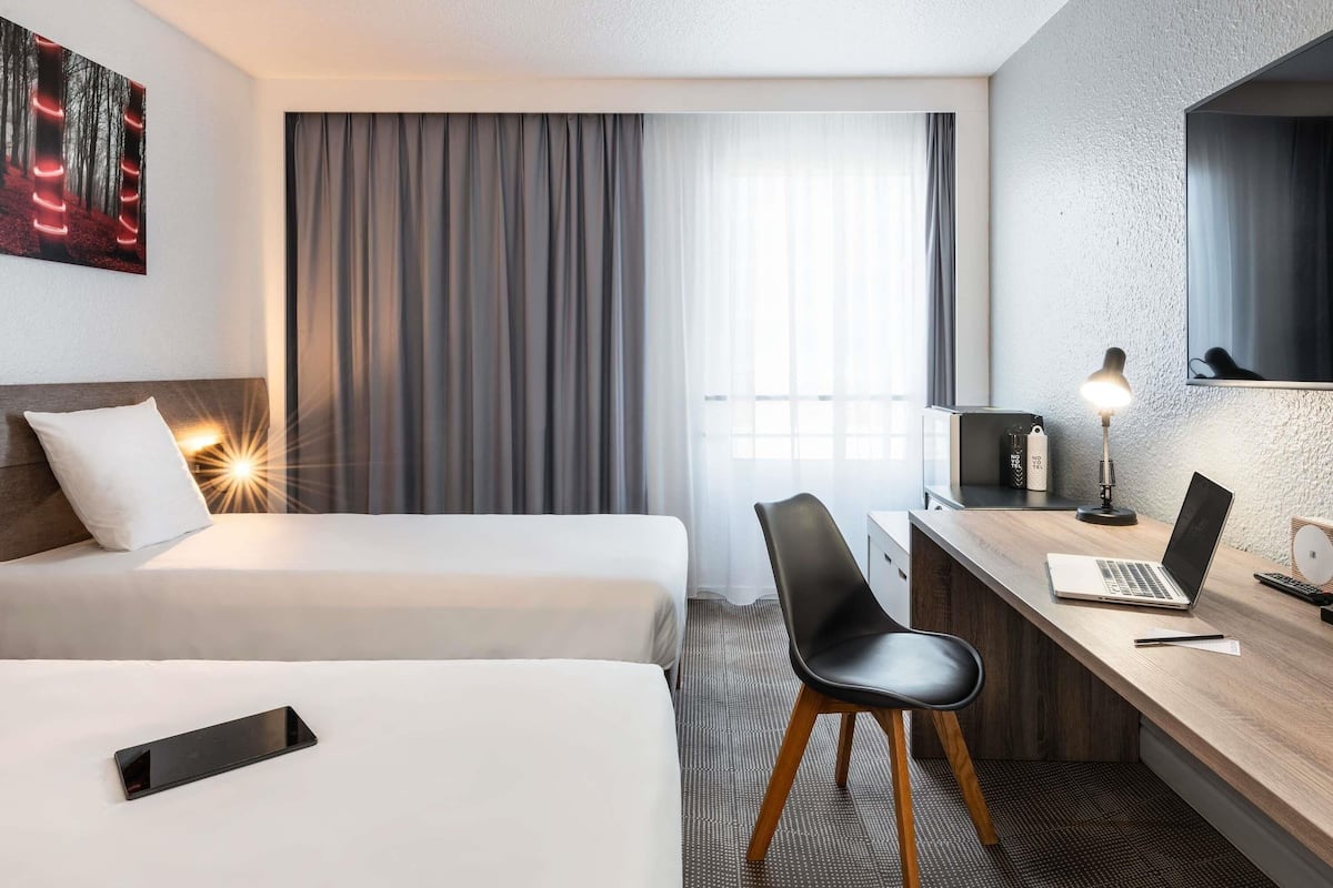 Habitación de hotel con dos camas, un escritorio con una computadora portátil, una silla, una lámpara de escritorio, un televisor y un cuadro en la pared.