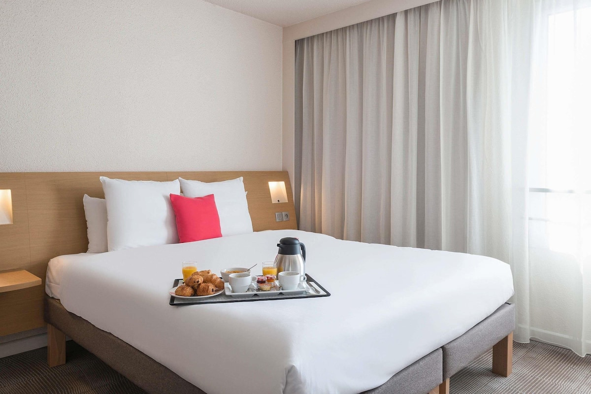 Una habitación de hotel con una cama bien hecha, una bandeja con alimentos para el desayuno, una mesita de noche y un amplio ventanal con cortinas.