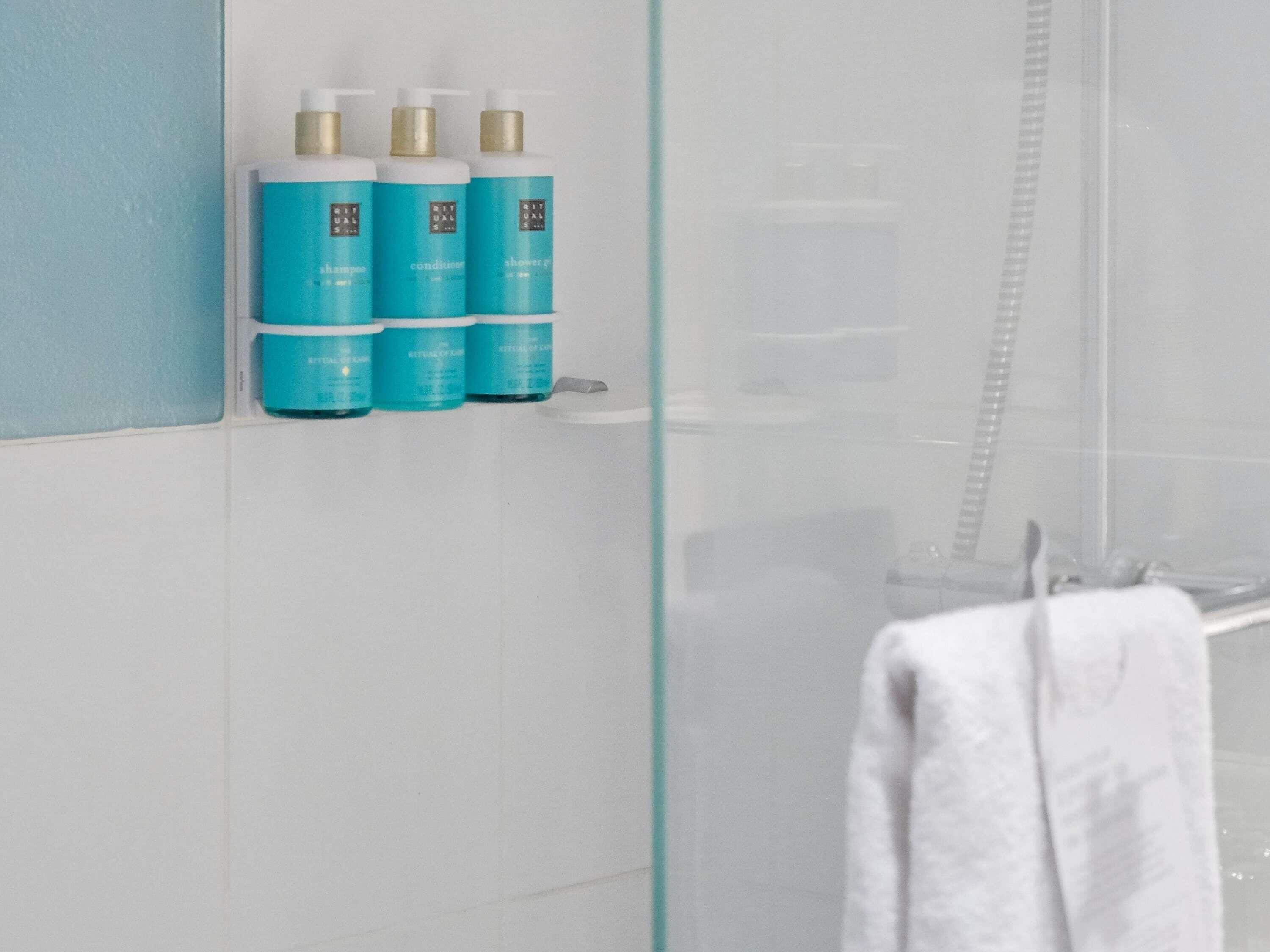 A bathroom with a glass shower enclosure, a white towel hanging on a rack, and three bottles of blue shampoo on a shelf.