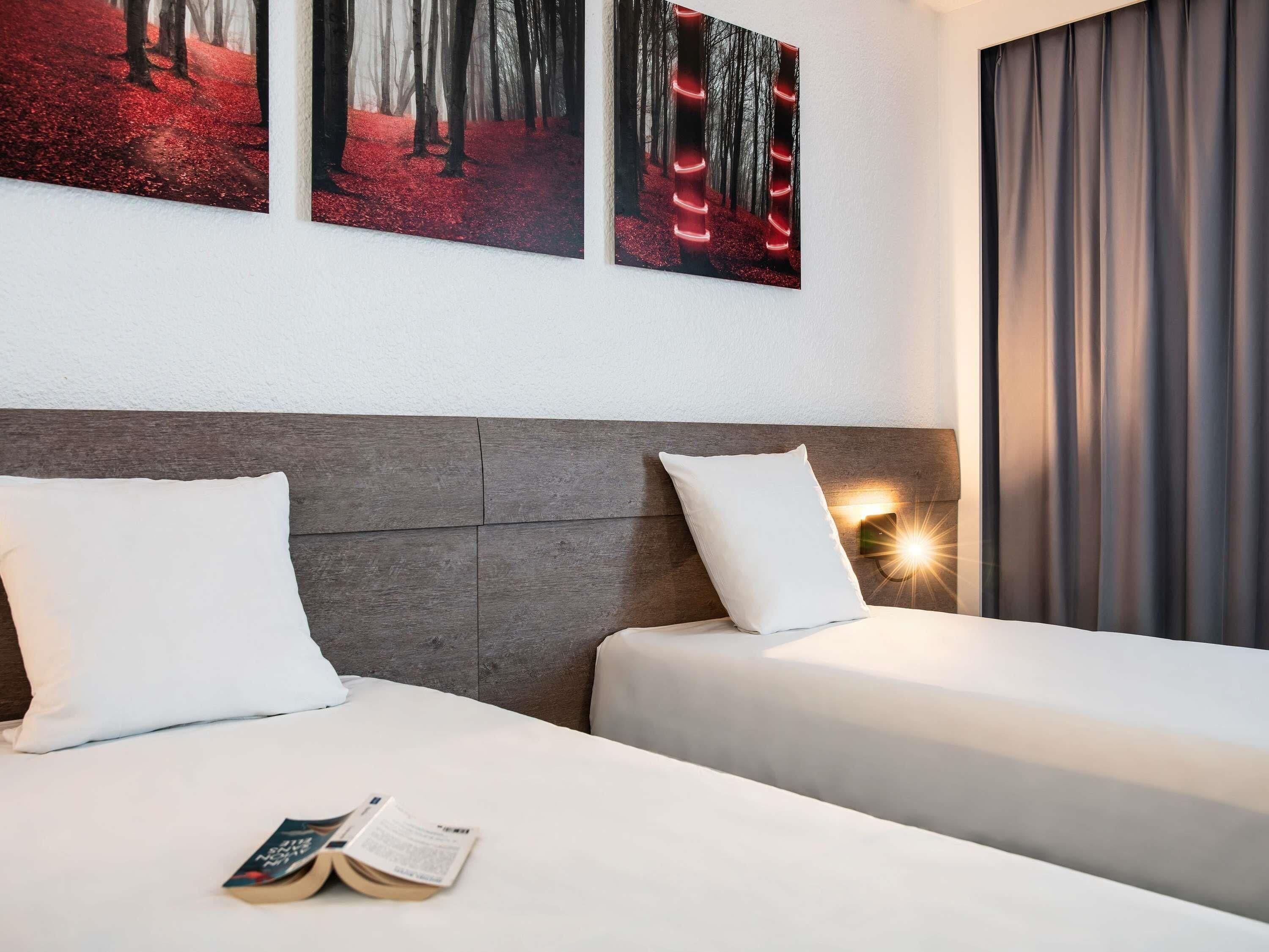 Hotelkamer met twee bedden, een houten hoofdbord, een nachtkastje met lamp en een groot raam met gordijnen.