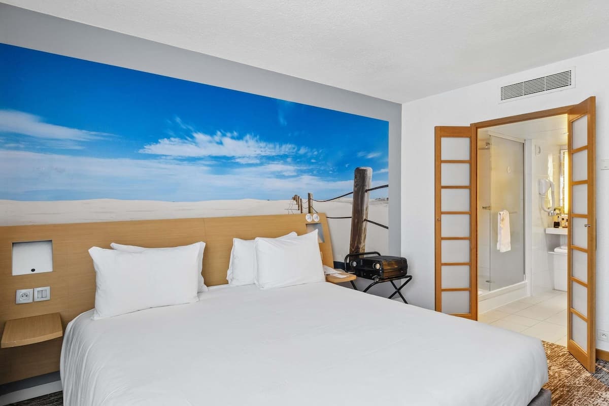 Habitación de hotel con una cama grande, cabecera de madera, un cuadro de un paisaje desértico, una puerta de madera que da a un baño y una mesita de noche con una lámpara.
