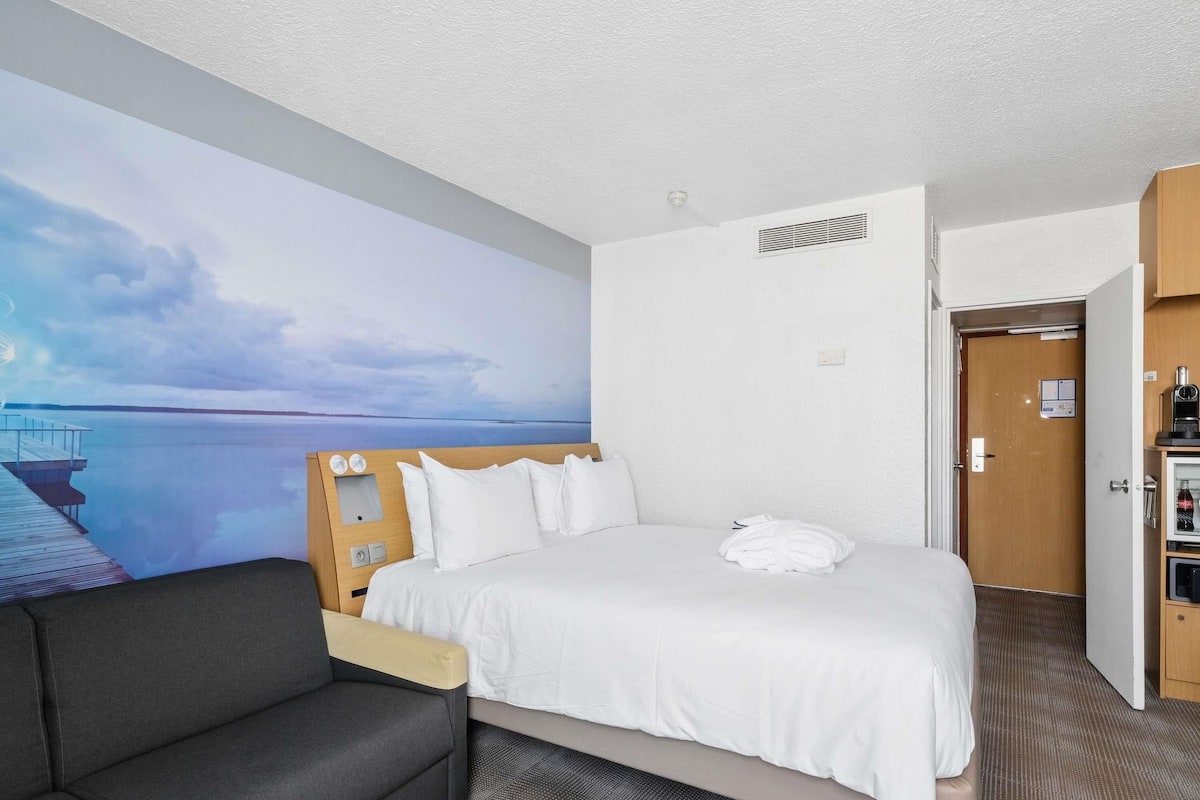 Habitación de hotel con cama, televisor montado en la pared, sofá, puerta y una pequeña kitchenette.