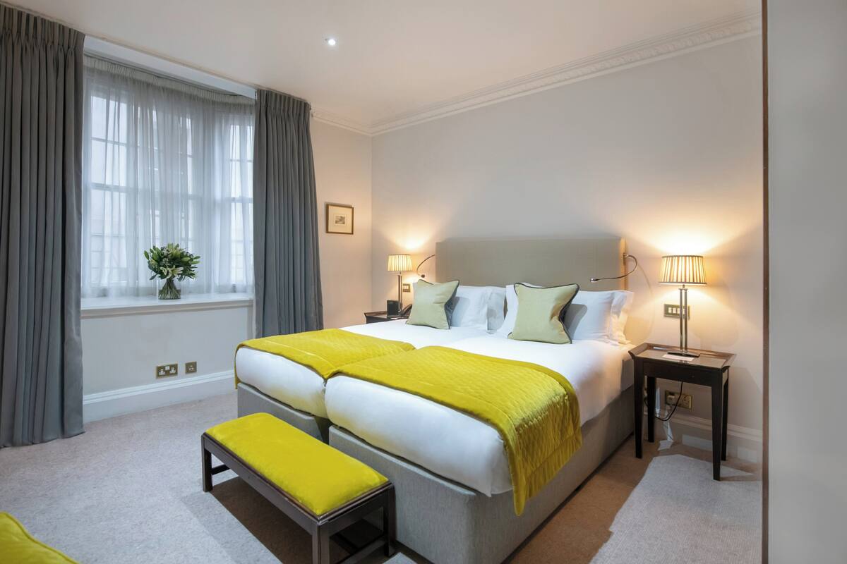 Une chambre d’hôtel avec un grand lit, deux tables de chevet, un banc, une fenêtre avec des rideaux et une lampe.