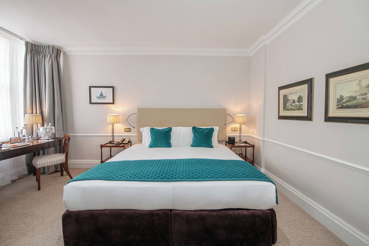 Une chambre d’hôtel avec un grand lit, deux tables de chevet avec des lampes, un bureau avec une chaise et des tableaux encadrés au mur.
