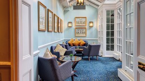 A hallway with a blue carpet, framed artwork on the walls, and a seating area with patterned chairs and cushions. - Dukes London (London)