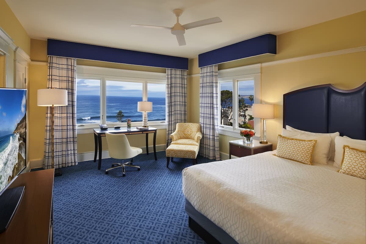 A hotel room with a bed, desk, chair, and a view of the ocean.