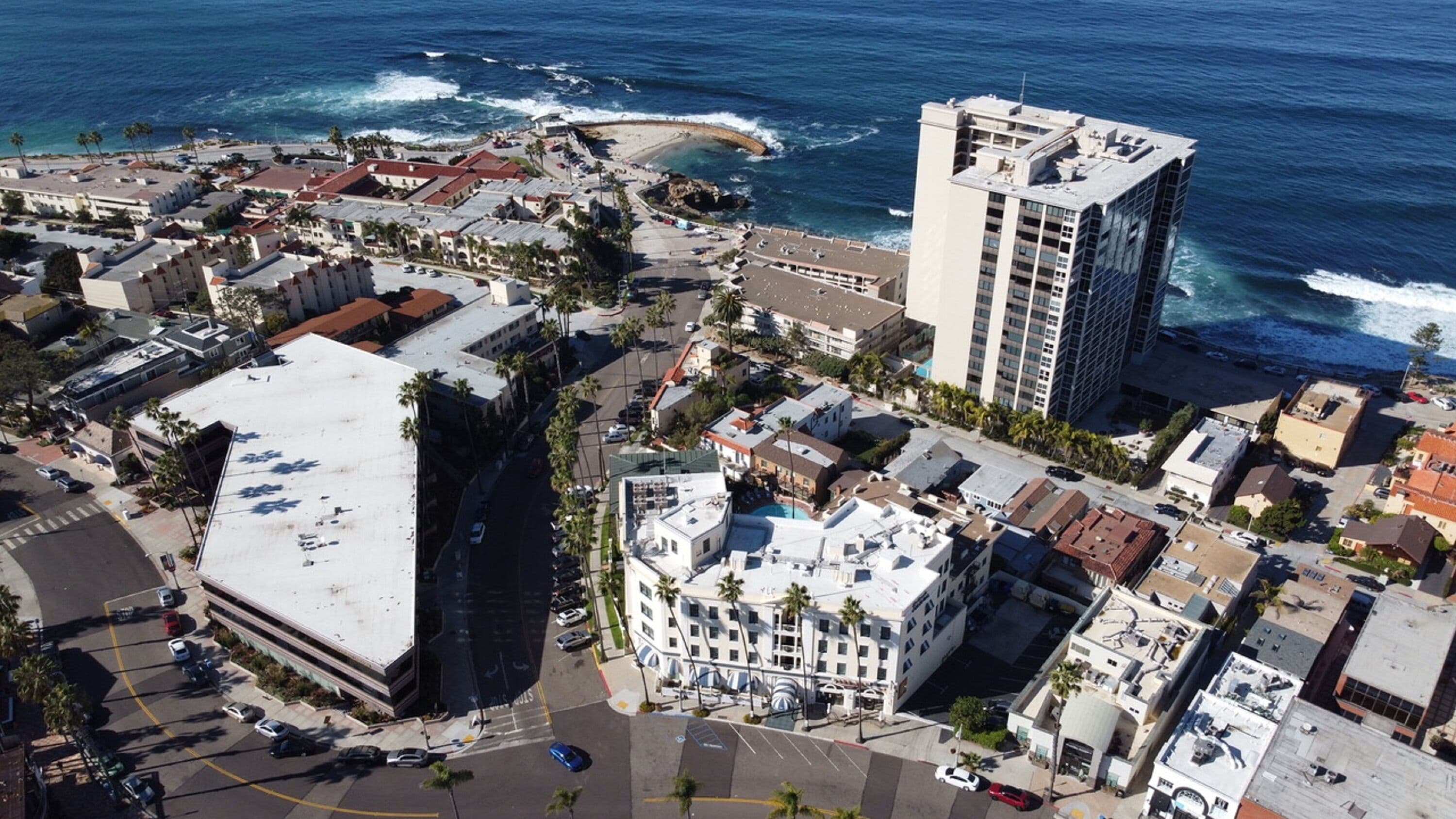 Vista aérea de una ciudad costera con un hotel destacado que cuenta con una piscina en la azotea, junto a una playa con un paseo curvo.