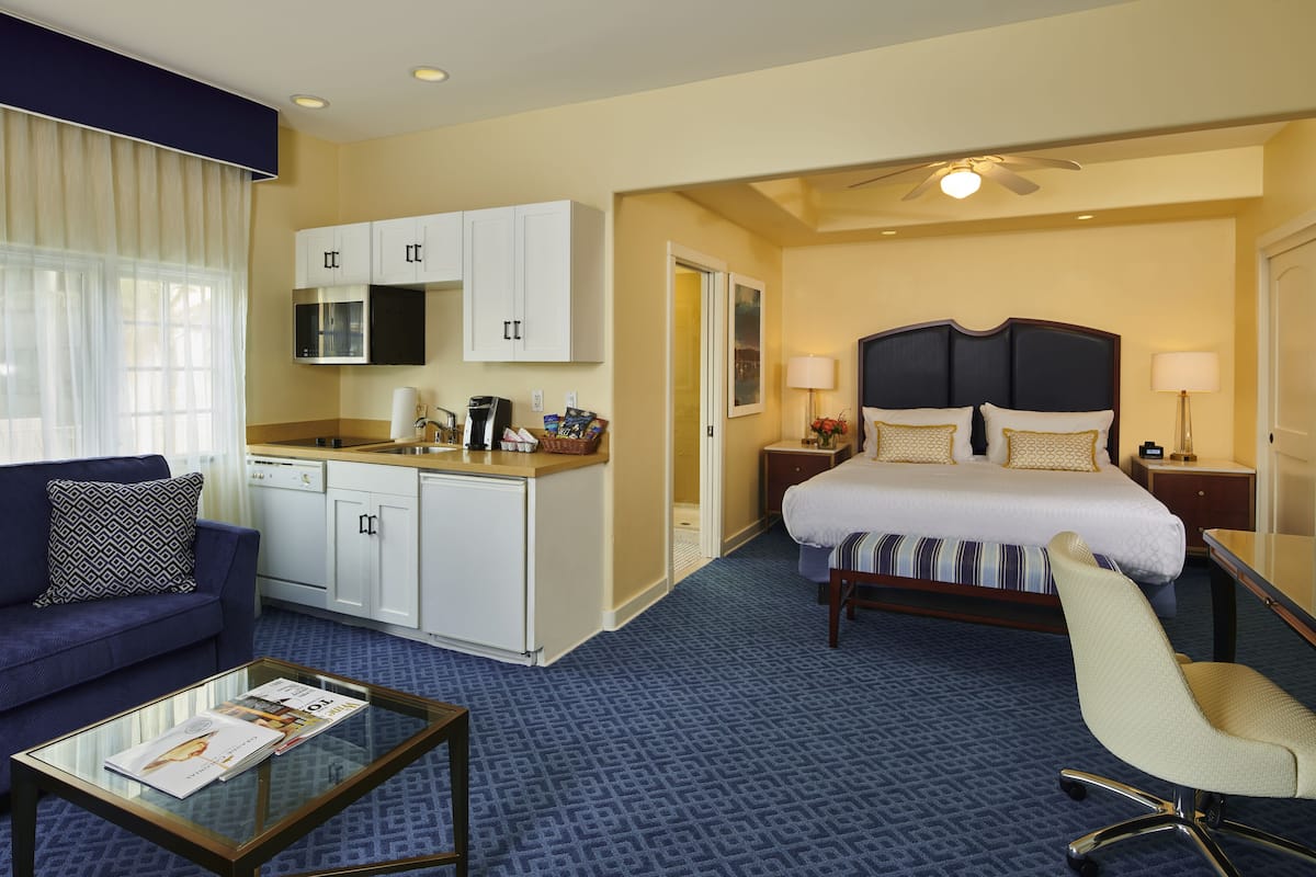 A hotel room with a bed, a sofa, a coffee table, a desk, a chair, a kitchenette, and a ceiling fan.