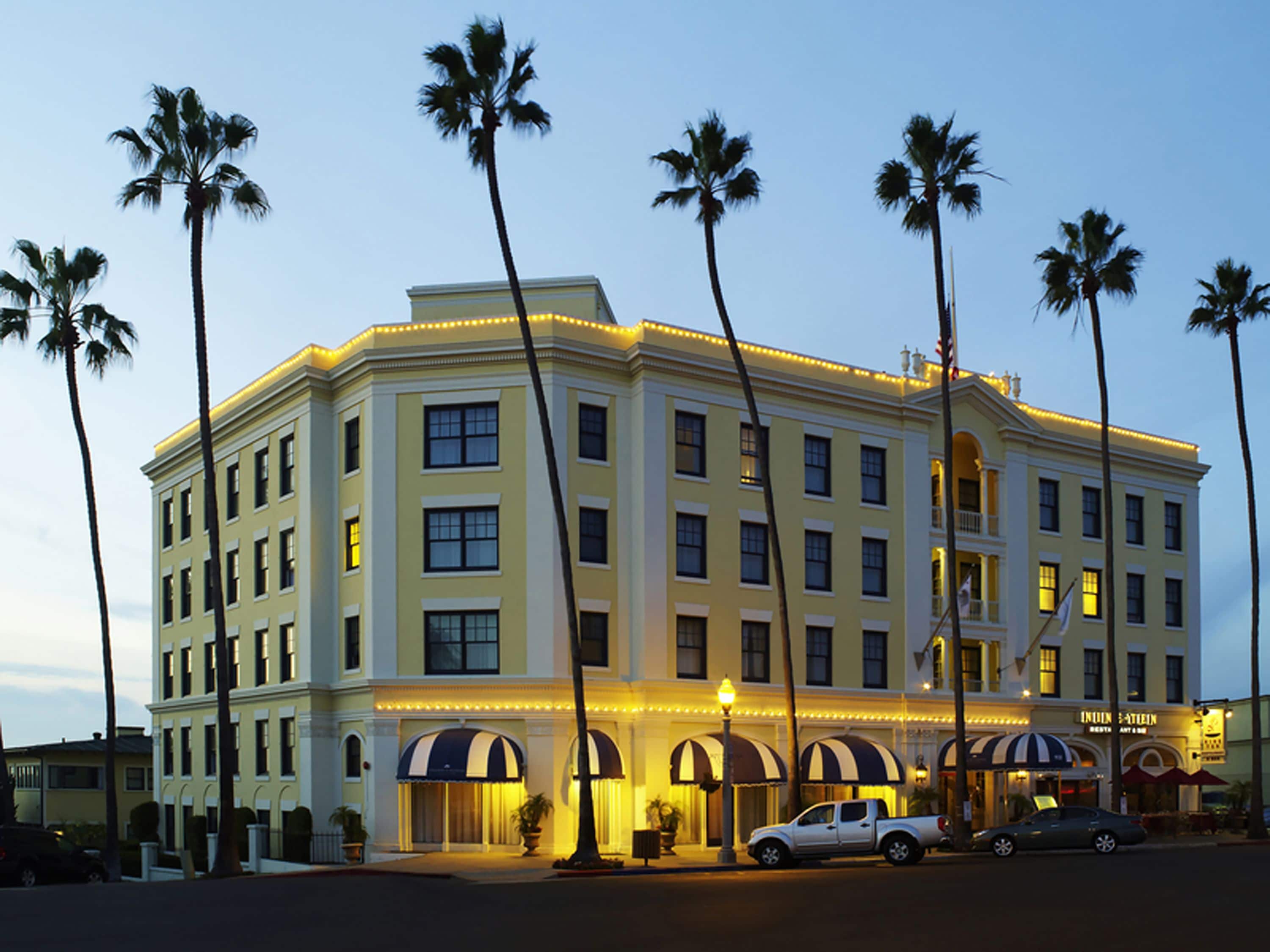 A multi-story hotel with a classic architectural style, featuring a central courtyard with palm trees, and a row of shops at the ground level.