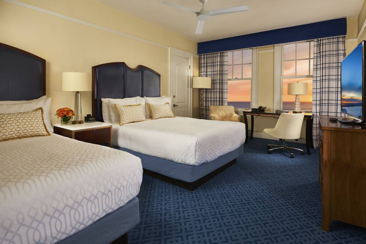 A hotel room with two beds, a desk, a chair, and a TV.