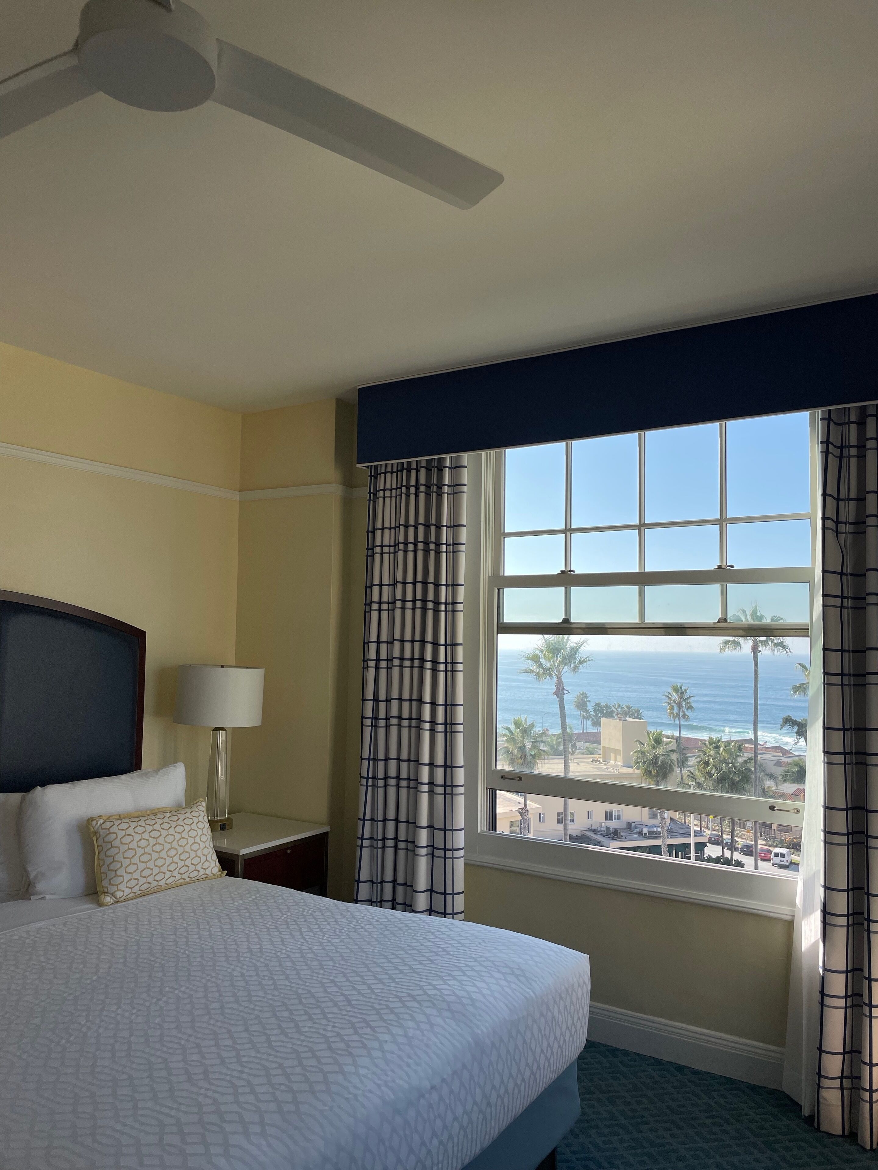 A hotel room with a bed, a nightstand with a lamp, a window with a view of the ocean and palm trees, and a ceiling fan.