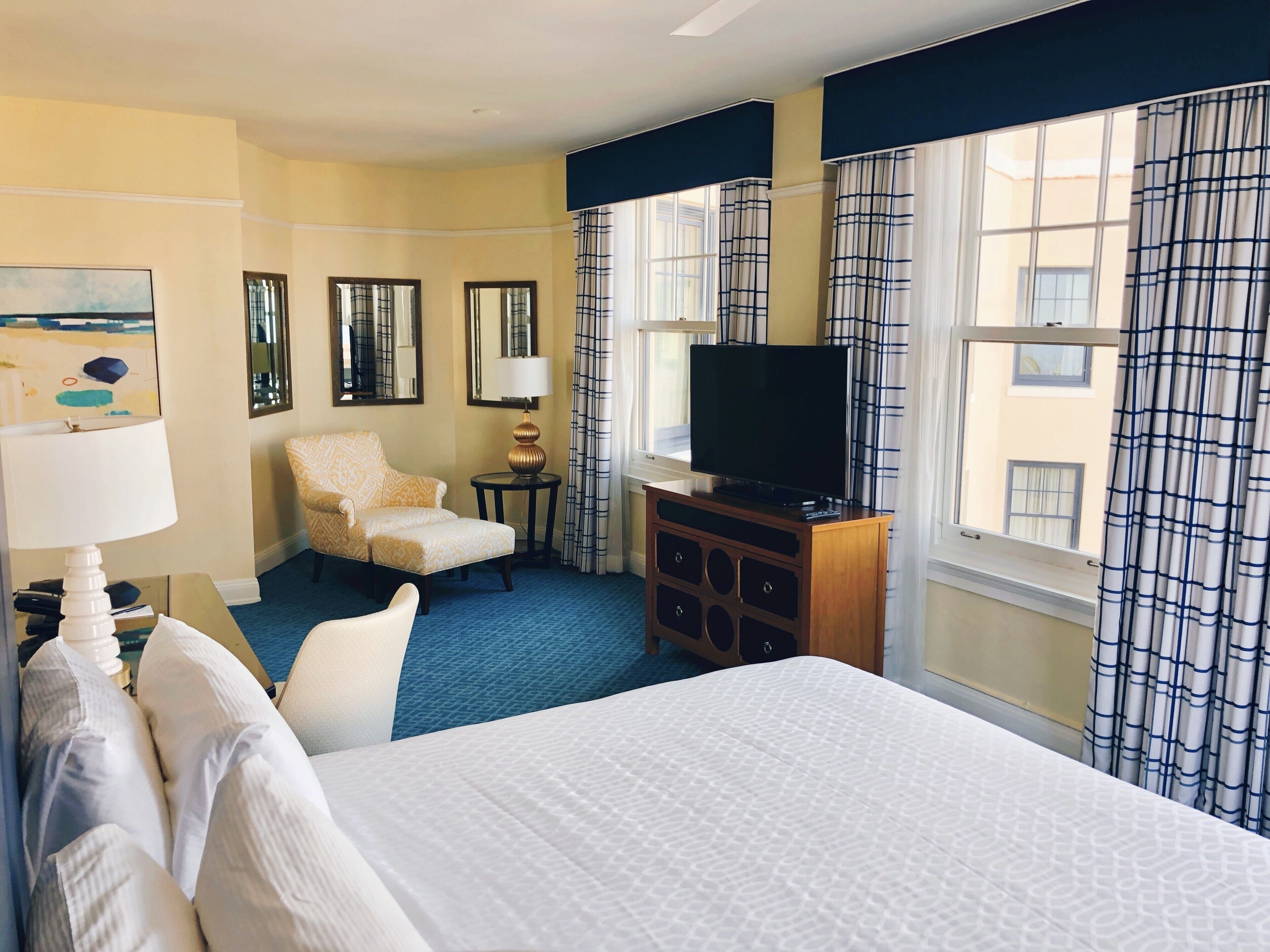 A hotel room with a bed, a TV on a wooden stand, a chair, a lamp, and a painting on the wall.