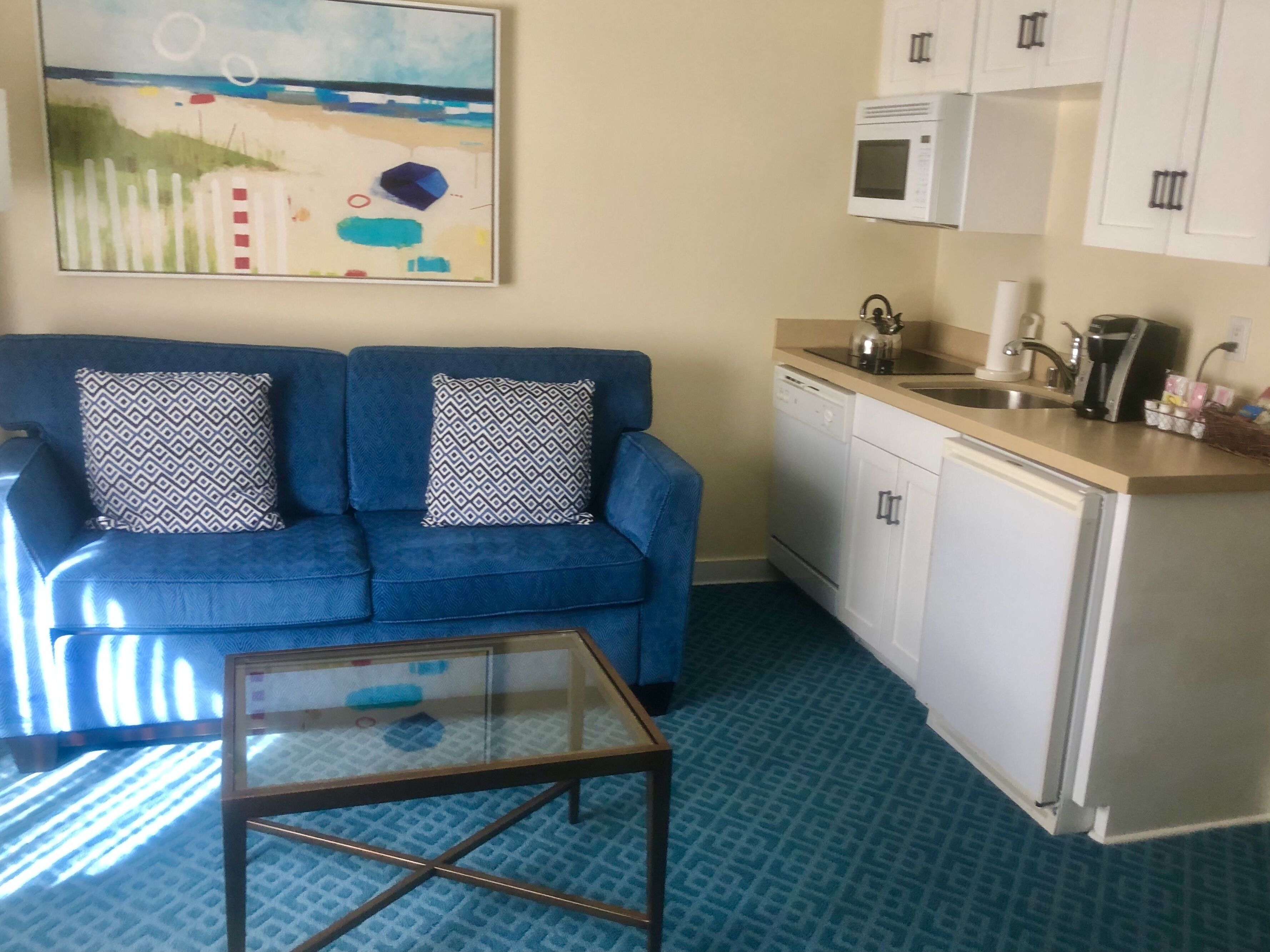 A compact living space with a blue sofa, patterned cushions, a glass coffee table, a kitchenette with a microwave, and a painting on the wall.