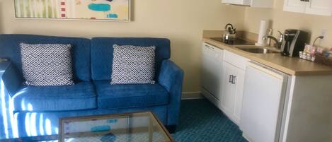 A compact living space with a blue sofa, patterned cushions, a glass coffee table, a kitchenette with a microwave, and a painting on the wall.