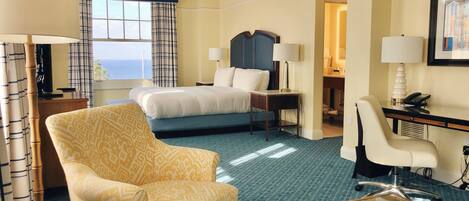 A hotel room with a bed, a desk, a chair, a glass coffee table, and a view of the ocean.