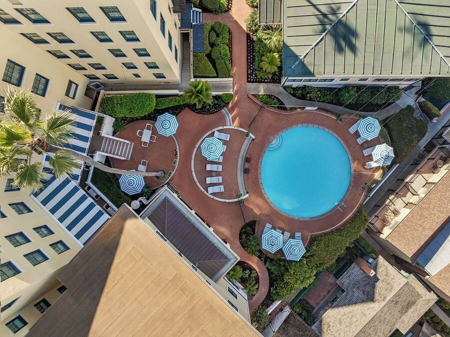 An aerial view of a hotel with a central swimming pool, multiple lounge areas with umbrellas, and a well-maintained garden.