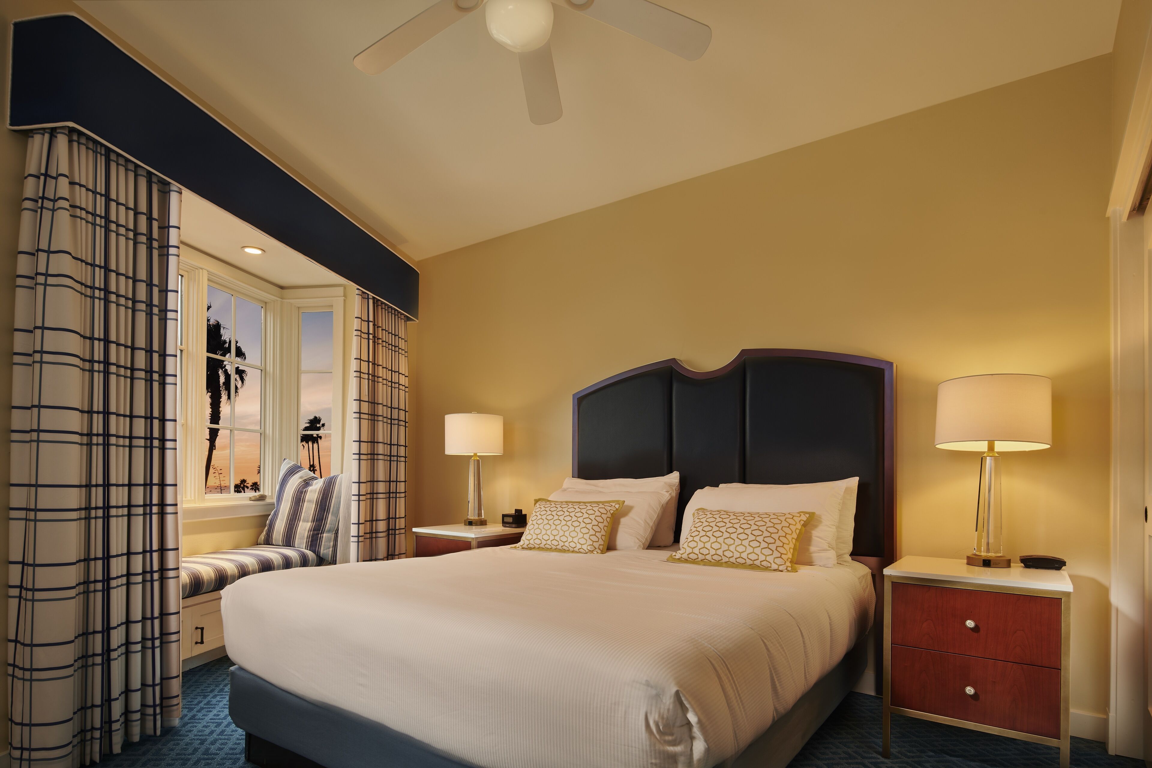 A hotel room with a large bed, two bedside tables with lamps, a window with curtains, and a ceiling fan.