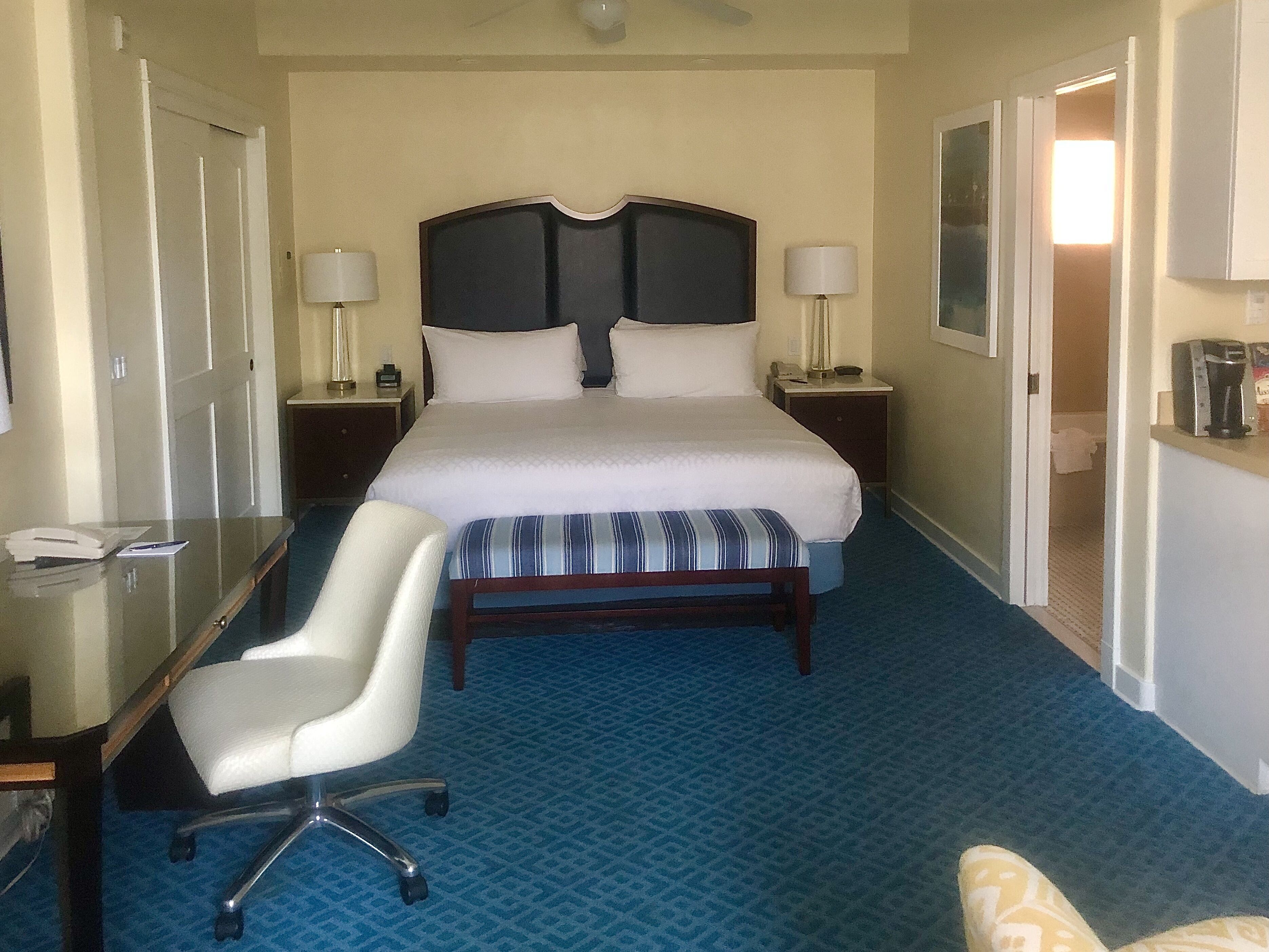 Habitación de hotel con una cama grande, un escritorio con silla, una mesita de noche con lámparas y un baño visible a través de una puerta abierta.