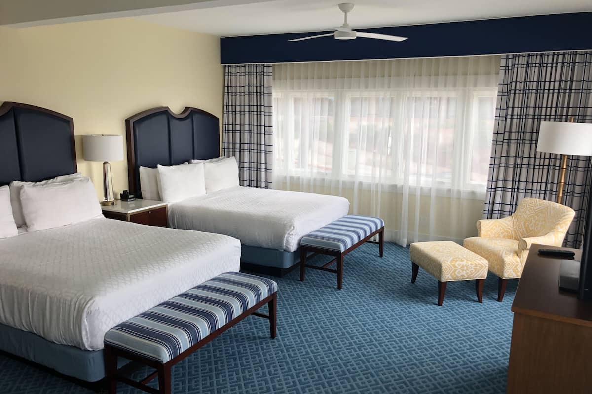 A hotel room with two beds, a bench, a chair, a desk, a lamp, and a ceiling fan.