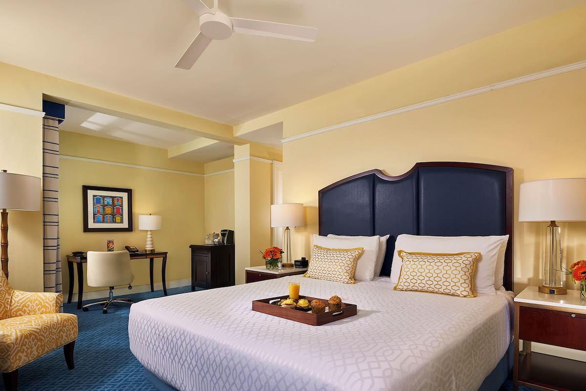 A hotel room with a large bed, a desk, a chair, a desk lamp, a floor lamp, and a ceiling fan.