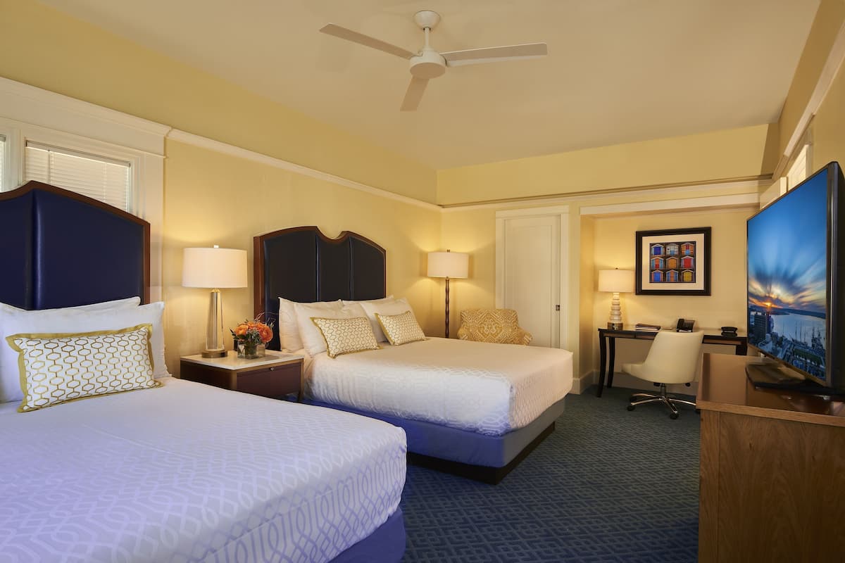 A hotel room with two beds, a desk, a chair, a television, and a ceiling fan.