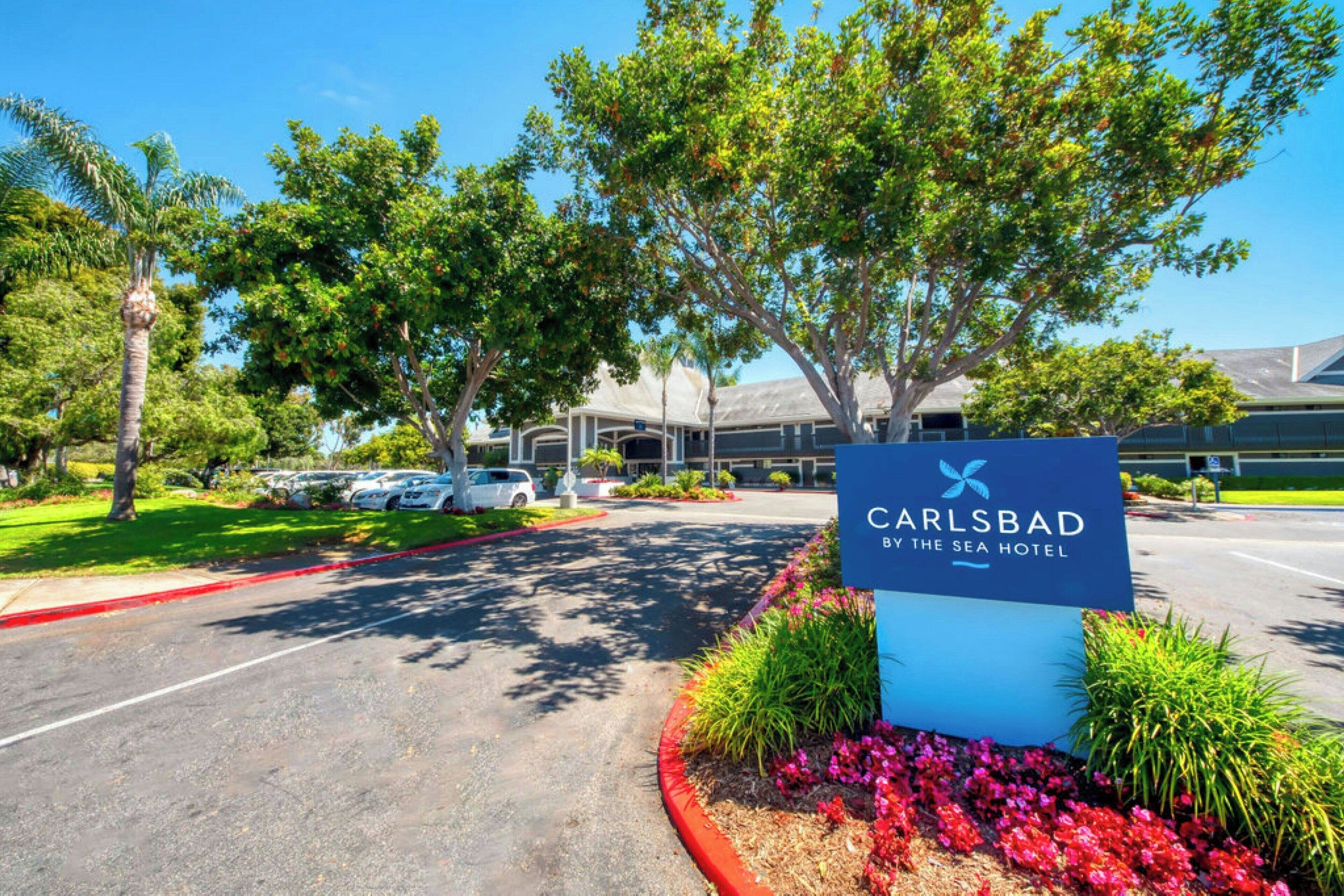 Entrada de un hotel con un letrero que dice 'Carlsbad by the Sea Hotel', rodeado de árboles y un estacionamiento.