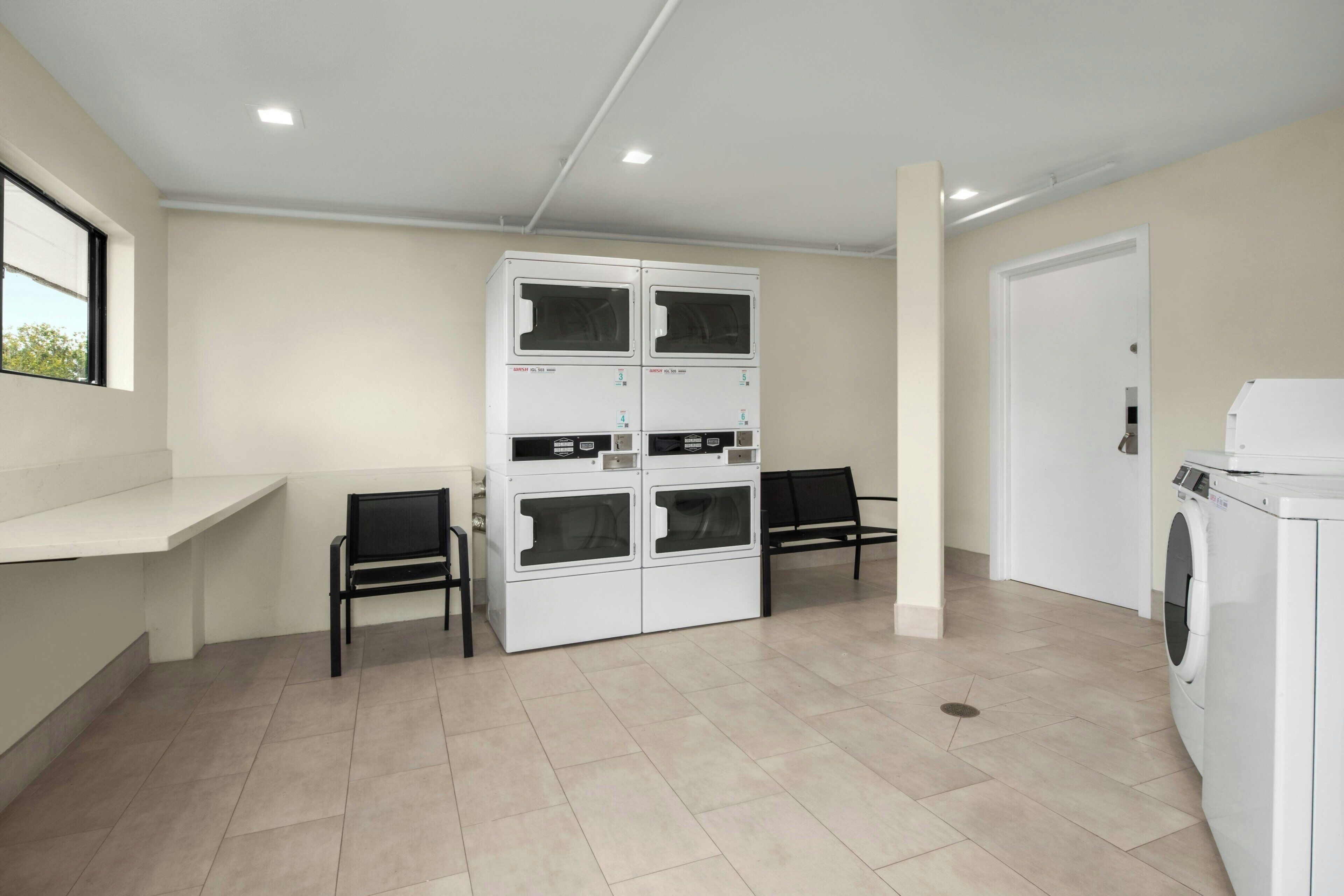 A laundry facility with a stacked washer and dryer unit, two chairs, a bench, and a door.