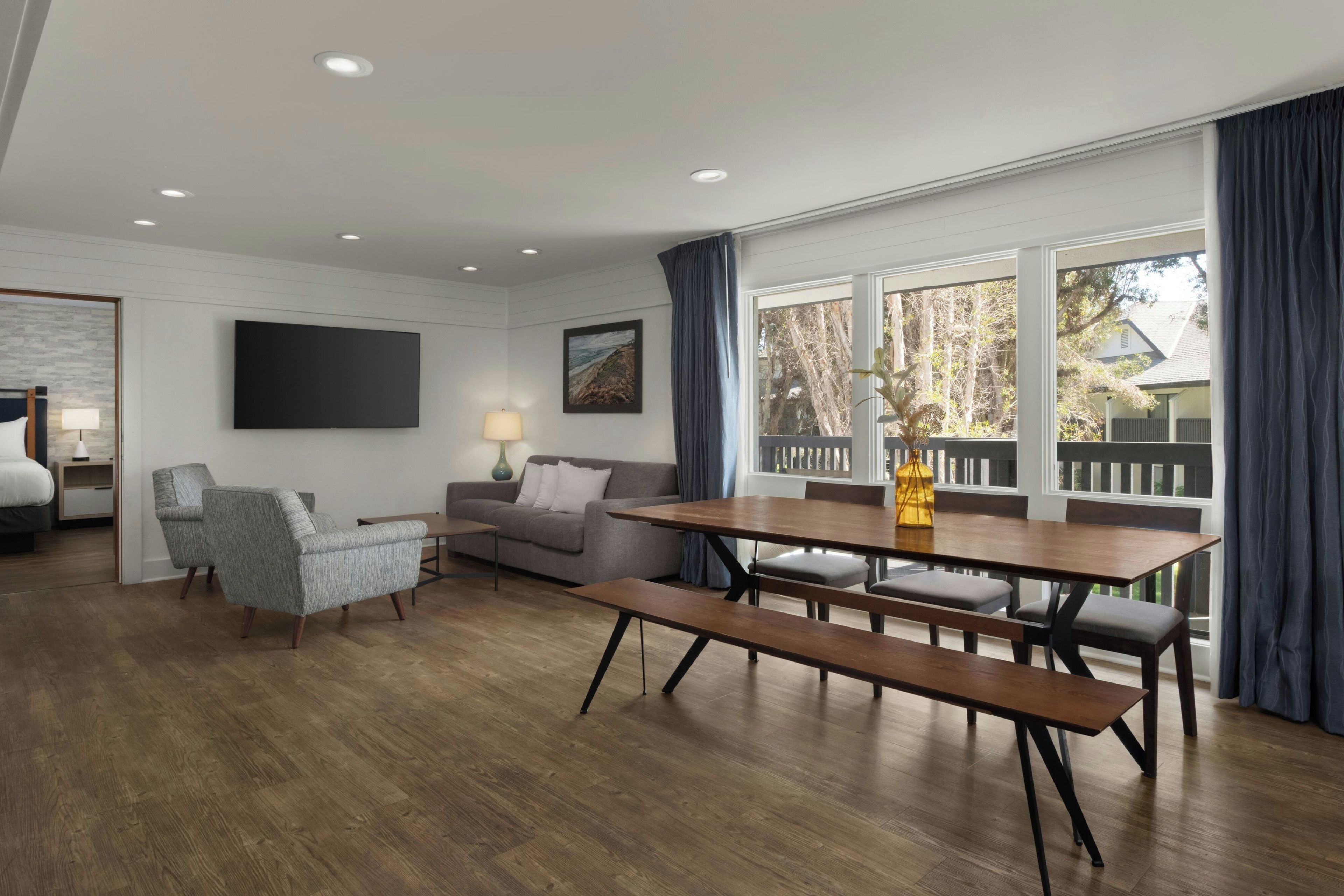 A modern living room with a sofa, a dining table with benches, a flat-screen TV mounted on the wall, and a large window with curtains.