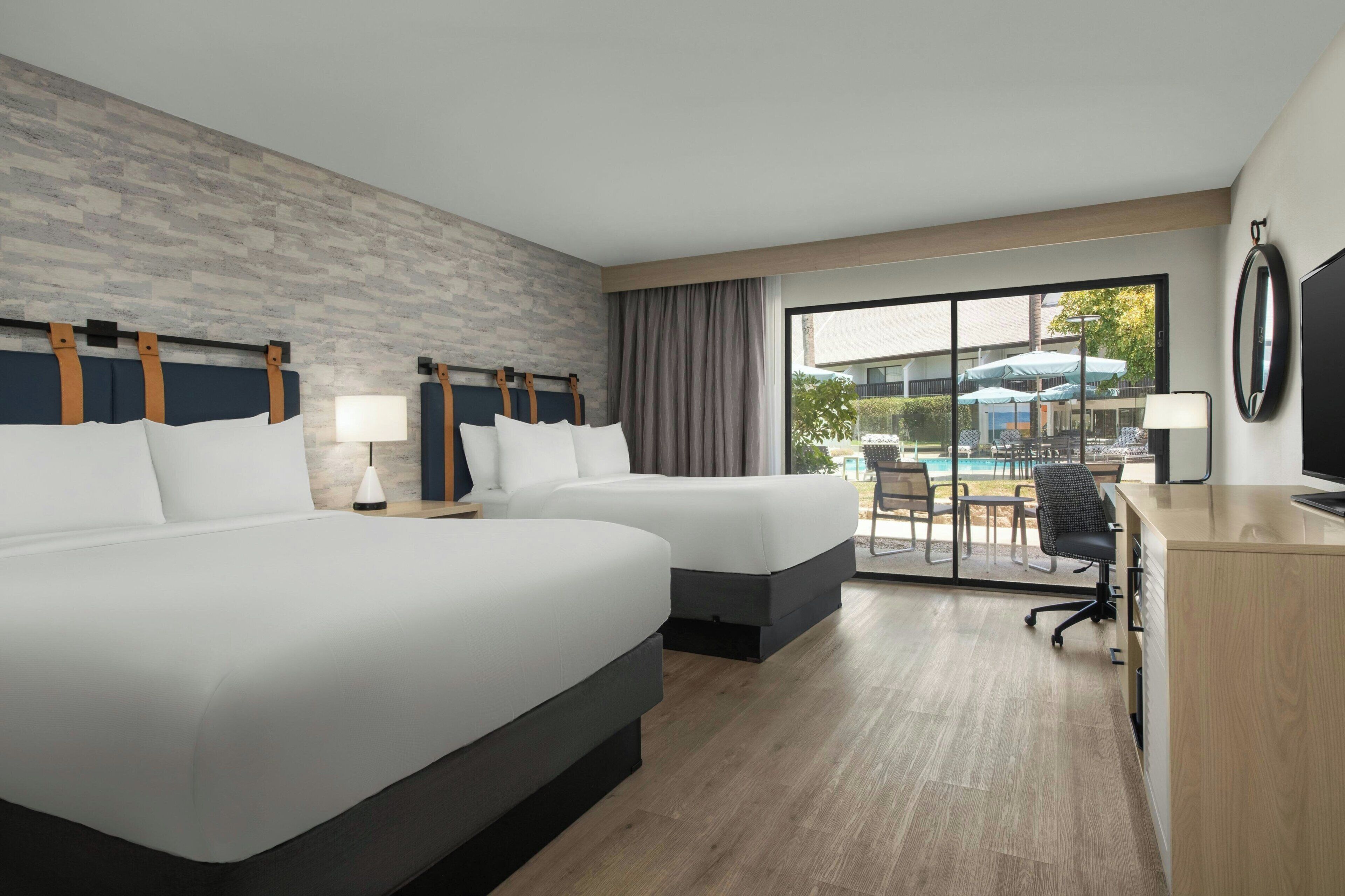 A hotel room with two beds, a desk, a chair, a TV, and a view of the outdoor pool area.
