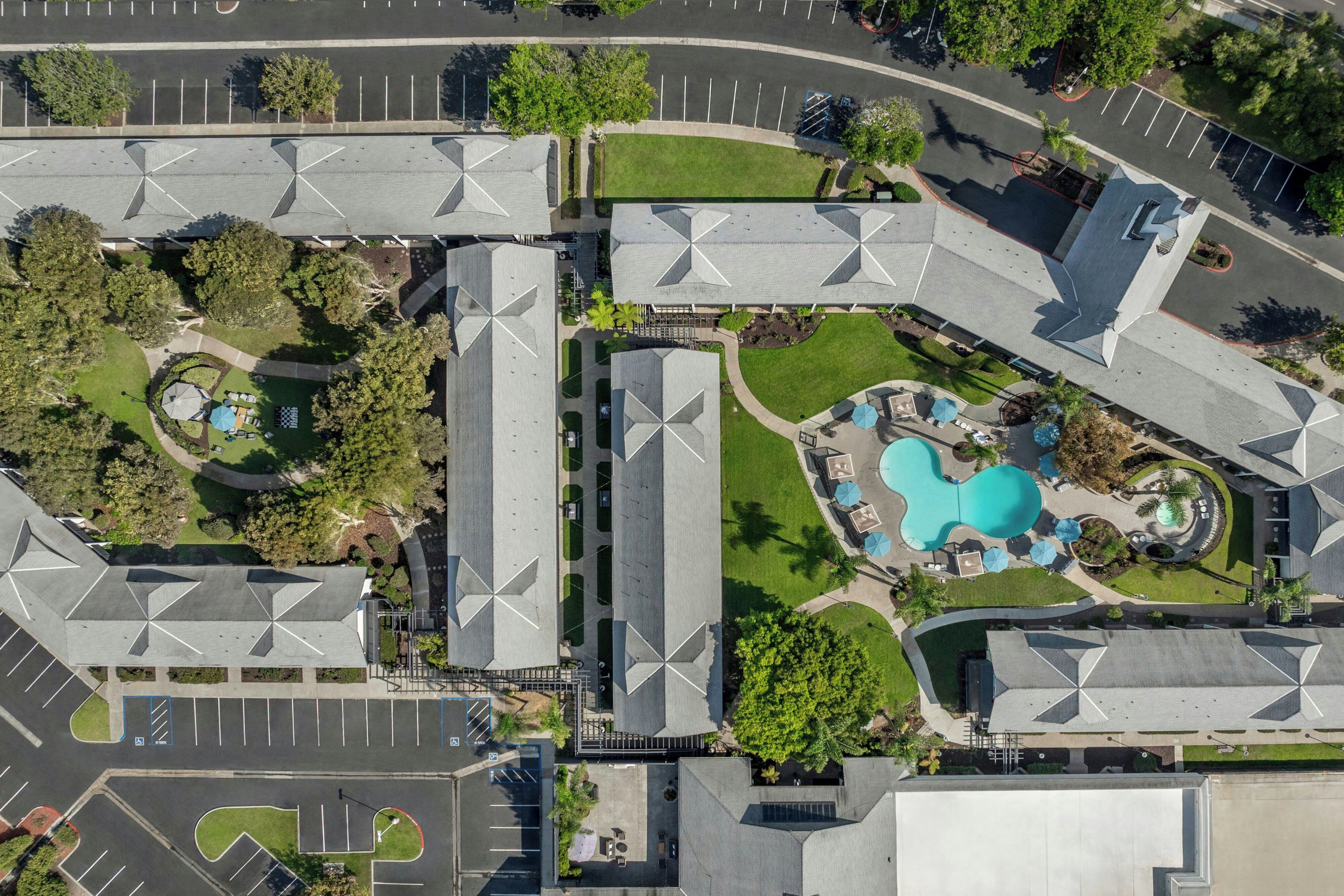 An aerial view of a modern building complex with multiple parking areas, a swimming pool, and landscaped gardens.