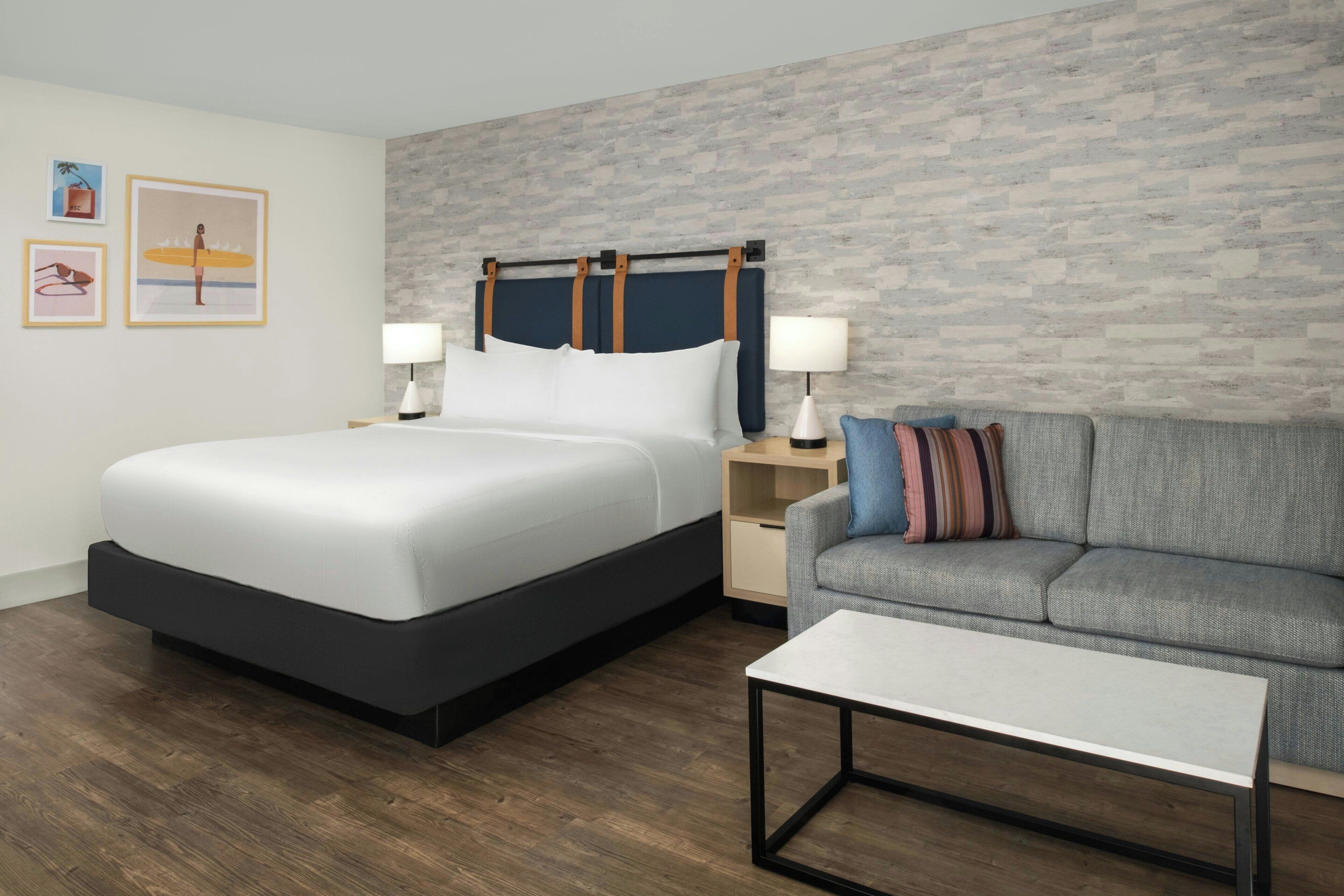 A modern hotel room with a large bed, a grey sofa, a small table, and a brick-patterned wall.